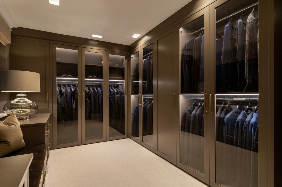 Large traditional walk-in wardrobe for men in Surrey with glass-front cabinets, brown cabinets, carpet and feature lighting.