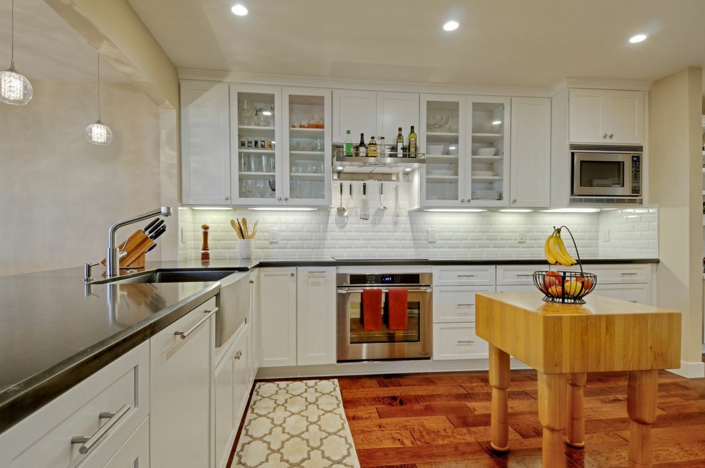 Example of a mid-sized transitional galley medium tone wood floor open concept kitchen design with a farmhouse sink, shaker cabinets, white cabinets, quartz countertops, white backsplash, ceramic backsplash, stainless steel appliances and no island