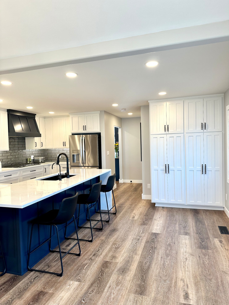 Mid-sized transitional l-shaped medium tone wood floor and brown floor open concept kitchen photo with an undermount sink, recessed-panel cabinets, white cabinets, quartz countertops, gray backsplash, ceramic backsplash, stainless steel appliances, an island and white countertops