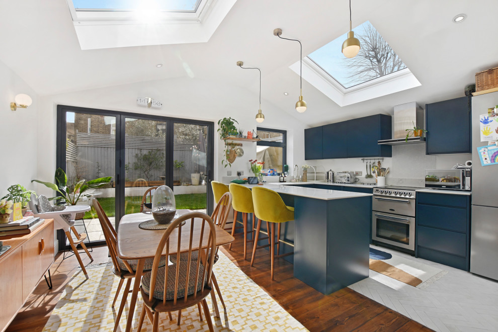 Mid-sized midcentury u-shaped eat-in kitchen in London with a drop-in sink, flat-panel cabinets, blue cabinets, solid surface benchtops, white splashback, ceramic splashback, stainless steel appliances, ceramic floors, a peninsula and white benchtop.
