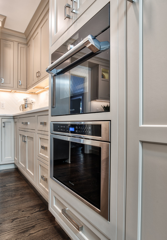 Mid-sized transitional l-shaped kitchen pantry in New York with a farmhouse sink, beaded inset cabinets, beige cabinets, granite benchtops, white splashback, porcelain splashback, stainless steel appliances, painted wood floors, with island, brown floor and white benchtop.