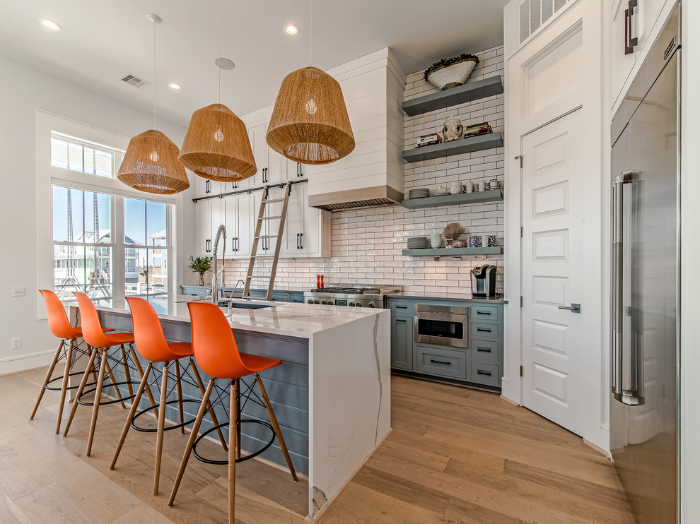 This is an example of a beach style l-shaped kitchen in Houston with an undermount sink, shaker cabinets, blue cabinets, white splashback, subway tile splashback, stainless steel appliances, light hardwood floors and with island.