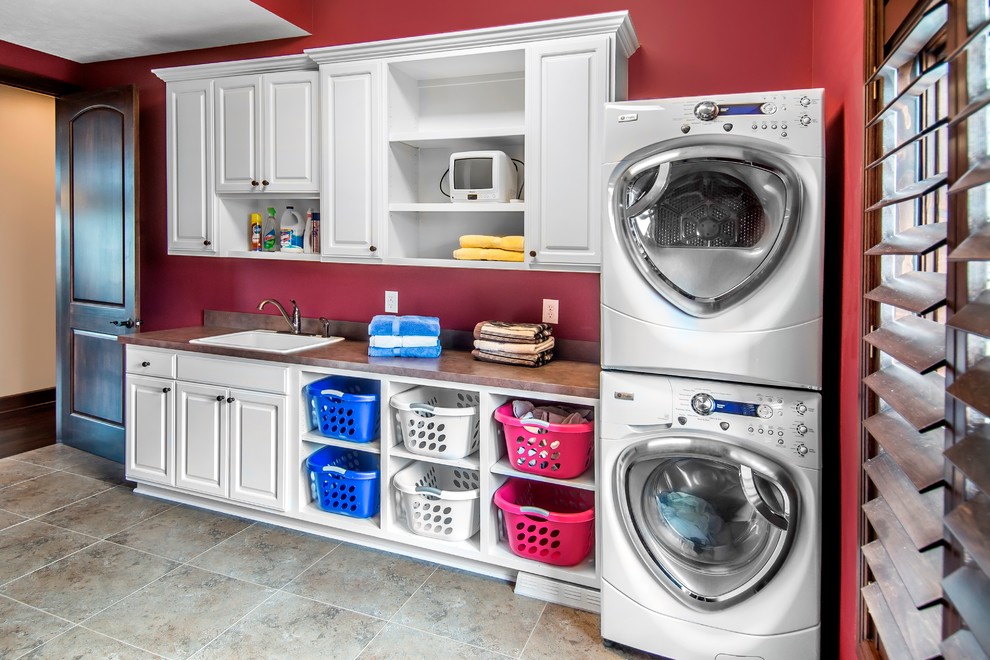 Design ideas for a mid-sized transitional single-wall dedicated laundry room in Omaha with raised-panel cabinets, white cabinets, laminate benchtops, red walls, porcelain floors, a stacked washer and dryer and a drop-in sink.
