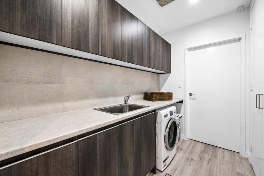 This is an example of a mid-sized beach style galley dedicated laundry room in Sunshine Coast with a drop-in sink, flat-panel cabinets, dark wood cabinets, quartz benchtops, beige walls, laminate floors, an integrated washer and dryer, brown floor and beige benchtop.