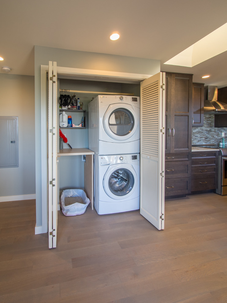 Photo of a small transitional single-wall laundry cupboard in Seattle with solid surface benchtops, medium hardwood floors, a stacked washer and dryer, grey walls, brown floor and beige benchtop.