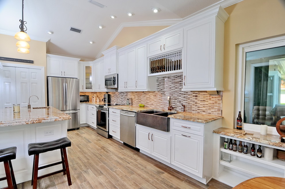 Example of a transitional medium tone wood floor and multicolored floor kitchen design in Miami with a farmhouse sink, raised-panel cabinets, white cabinets, granite countertops, multicolored backsplash, ceramic backsplash, stainless steel appliances and an island