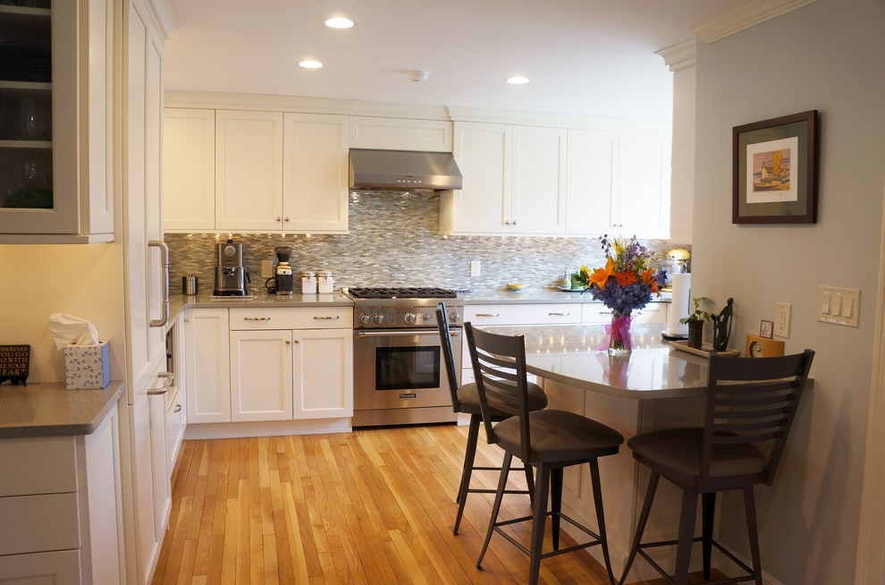 Example of a mid-sized transitional l-shaped light wood floor eat-in kitchen design in Boston with a farmhouse sink, shaker cabinets, white cabinets, quartz countertops, blue backsplash, glass tile backsplash, paneled appliances and a peninsula