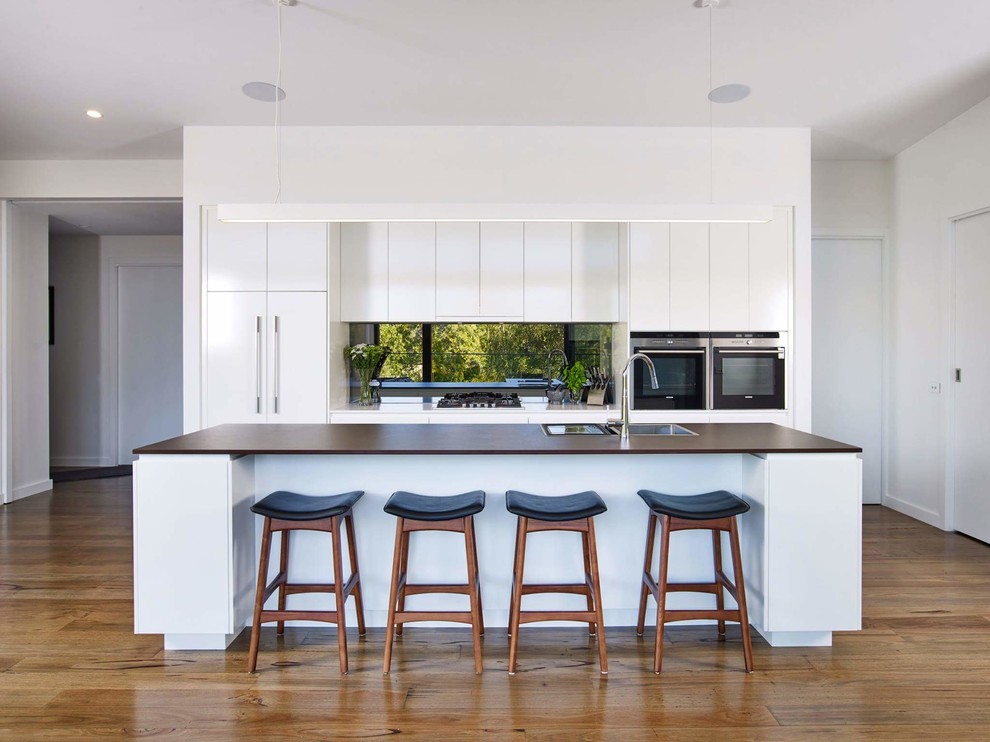 This is an example of a contemporary galley kitchen pantry in Other with a drop-in sink, flat-panel cabinets, white cabinets, solid surface benchtops, metallic splashback, mirror splashback, stainless steel appliances, medium hardwood floors, with island, brown floor and brown benchtop.