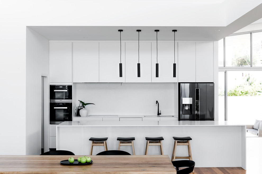 This is an example of a contemporary galley kitchen in Sunshine Coast with a double-bowl sink, flat-panel cabinets, white cabinets, white splashback, black appliances, dark hardwood floors, with island and brown floor.