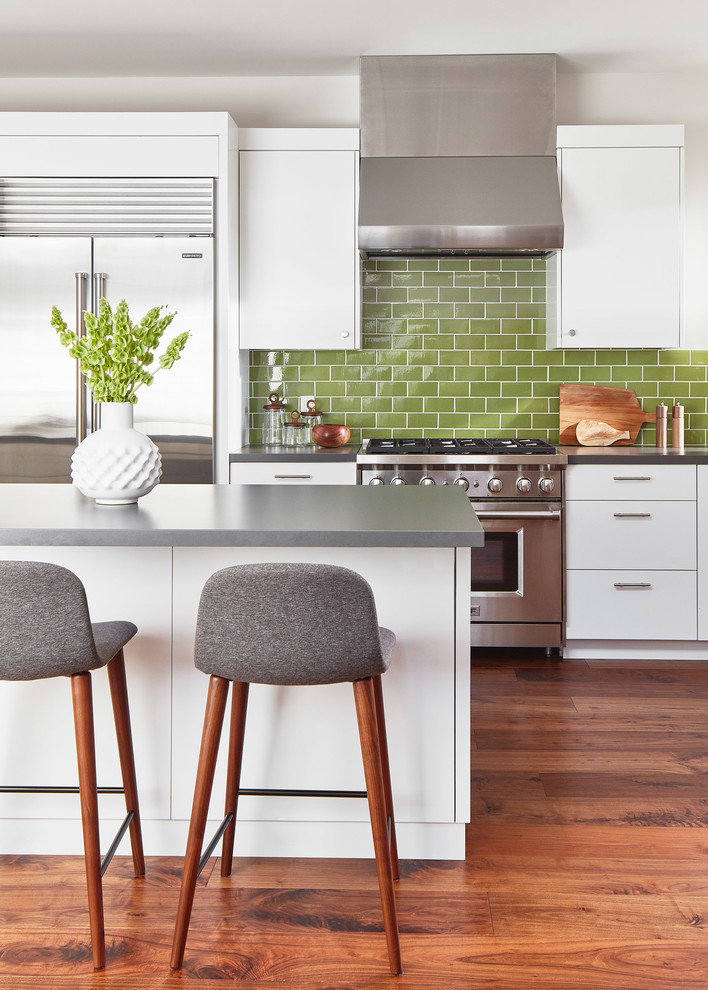 Photo of a contemporary galley kitchen in San Francisco with flat-panel cabinets, white cabinets, green splashback, subway tile splashback, stainless steel appliances, medium hardwood floors, with island, brown floor and grey benchtop.