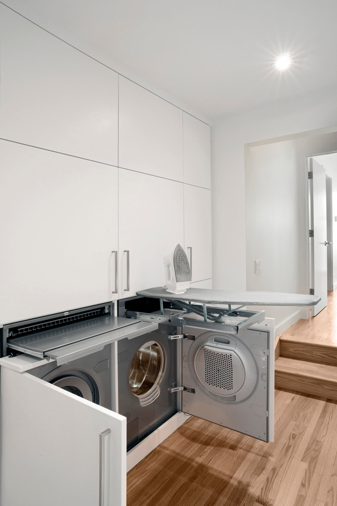 Design ideas for a small contemporary laundry cupboard in Calgary with flat-panel cabinets, white cabinets, white walls, medium hardwood floors and a side-by-side washer and dryer.