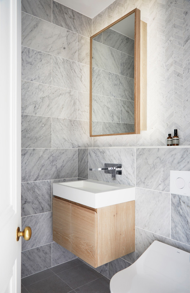 Пример оригинального дизайна: маленькая ванная комната в стиле модернизм с плоскими фасадами, светлыми деревянными фасадами, ванной в нише, душем над ванной, инсталляцией, серой плиткой, мраморной плиткой, серыми стенами, полом из керамогранита, подвесной раковиной, серым полом, шторкой для ванной, тумбой под одну раковину и подвесной тумбой для на участке и в саду