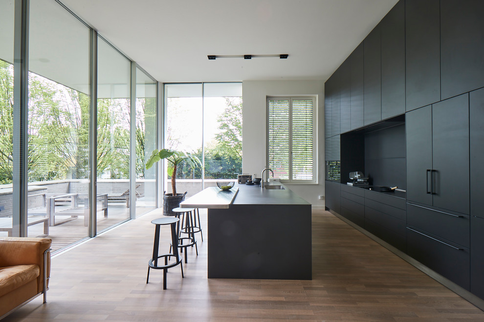 Large modern galley eat-in kitchen in Hamburg with flat-panel cabinets, black cabinets, black splashback, black appliances, with island, black benchtop, a single-bowl sink, concrete benchtops, medium hardwood floors and brown floor.