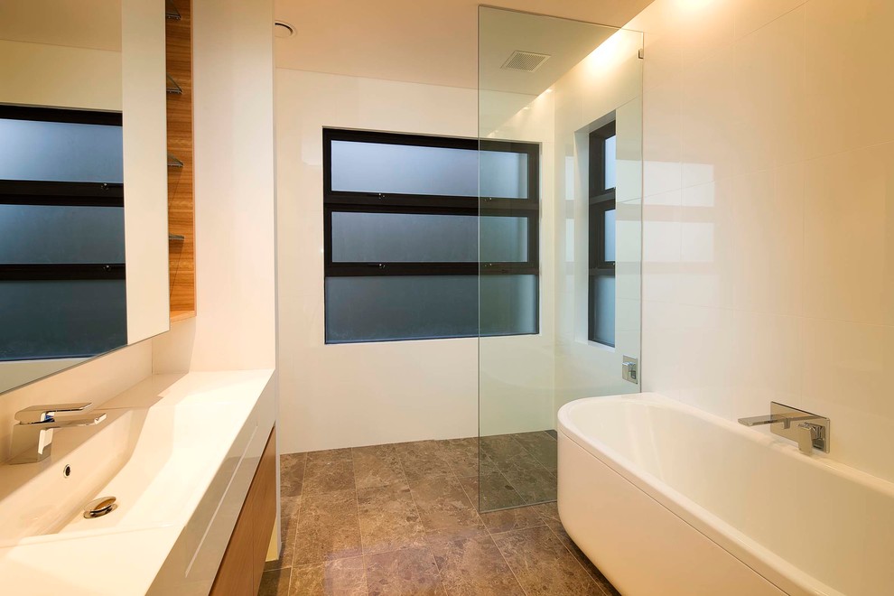 This is an example of a contemporary master bathroom in Sydney with an integrated sink, flat-panel cabinets, medium wood cabinets, solid surface benchtops, a freestanding tub, a corner shower, brown tile, white walls and travertine floors.