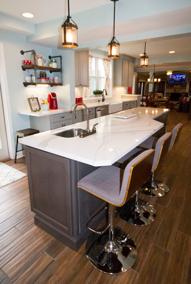 Example of a large transitional l-shaped medium tone wood floor and brown floor enclosed kitchen design in Cincinnati with a farmhouse sink, recessed-panel cabinets, gray cabinets, marble countertops, white backsplash, porcelain backsplash, stainless steel appliances and an island