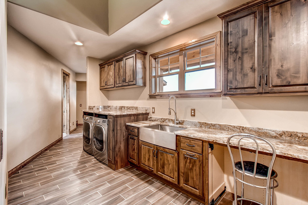 Large country single-wall laundry room in Denver with a farmhouse sink, raised-panel cabinets, dark wood cabinets, granite benchtops, beige walls, porcelain floors, a side-by-side washer and dryer, brown floor and multi-coloured benchtop.