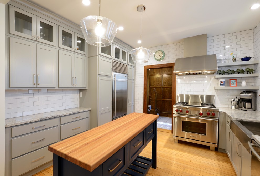 Mid-sized transitional light wood floor and brown floor enclosed kitchen photo with a farmhouse sink, shaker cabinets, gray cabinets, wood countertops, white backsplash, porcelain backsplash, stainless steel appliances, an island and brown countertops