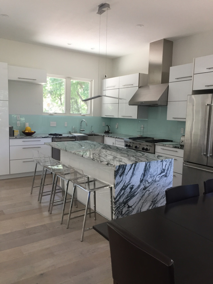 Inspiration for a large modern l-shaped eat-in kitchen in Minneapolis with a farmhouse sink, flat-panel cabinets, white cabinets, granite benchtops, green splashback, glass sheet splashback, stainless steel appliances, light hardwood floors, with island and multi-coloured benchtop.