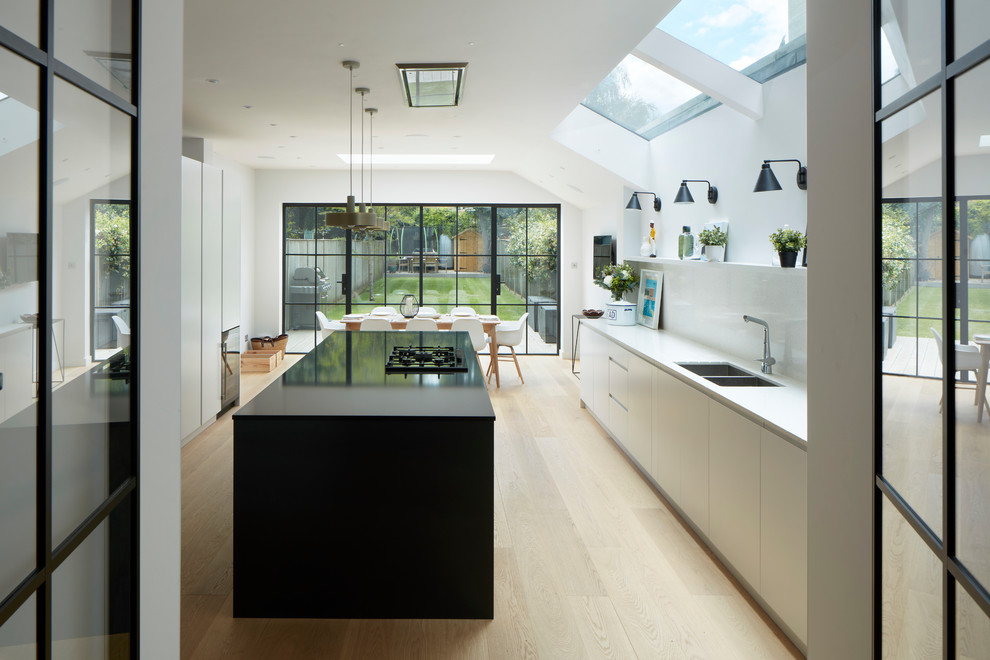 Design ideas for a transitional galley eat-in kitchen in London with an undermount sink, flat-panel cabinets, black cabinets, white splashback, panelled appliances, light hardwood floors, with island, beige floor and black benchtop.