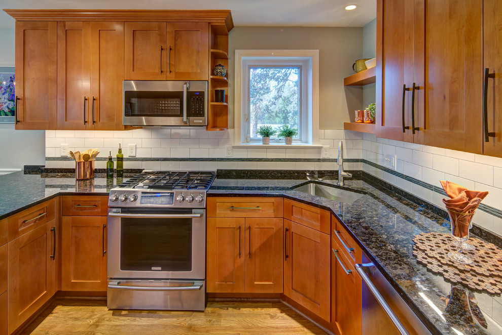 This is an example of a mid-sized transitional u-shaped eat-in kitchen in DC Metro with an undermount sink, shaker cabinets, medium wood cabinets, quartz benchtops, white splashback, porcelain splashback, stainless steel appliances, light hardwood floors, a peninsula, brown floor and black benchtop.