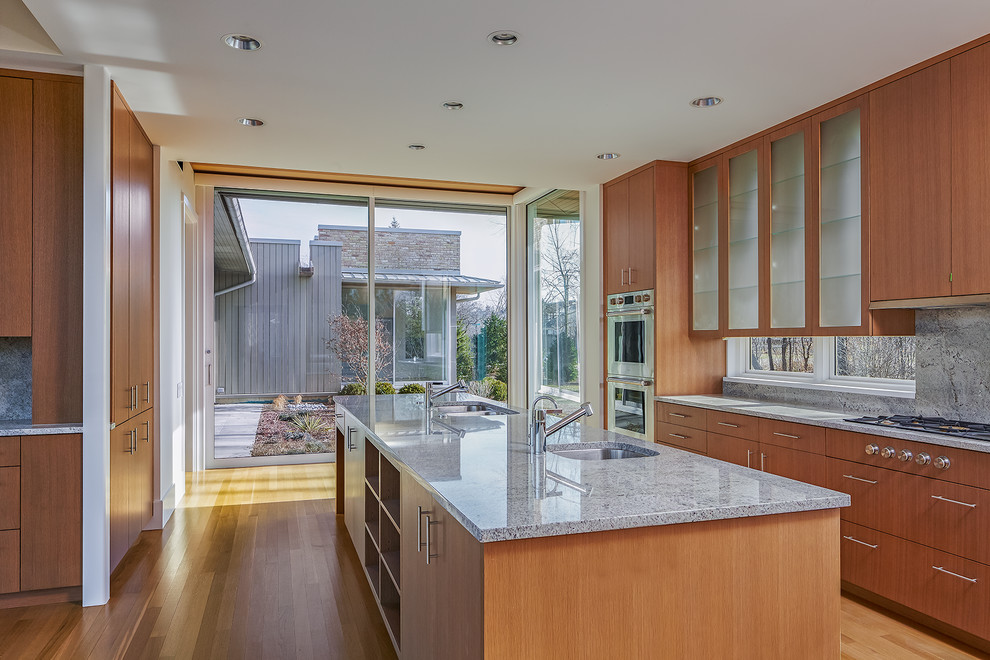 Large modern u-shaped eat-in kitchen in Chicago with a double-bowl sink, flat-panel cabinets, medium wood cabinets, limestone benchtops, grey splashback, limestone splashback, stainless steel appliances, with island, grey benchtop and light hardwood floors.