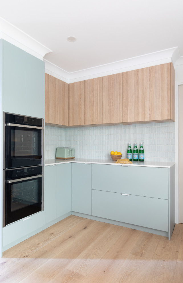 Design ideas for a mid-sized beach style l-shaped kitchen pantry in Brisbane with an undermount sink, flat-panel cabinets, green cabinets, quartz benchtops, blue splashback, mosaic tile splashback, stainless steel appliances, light hardwood floors, a peninsula, beige floor and white benchtop.
