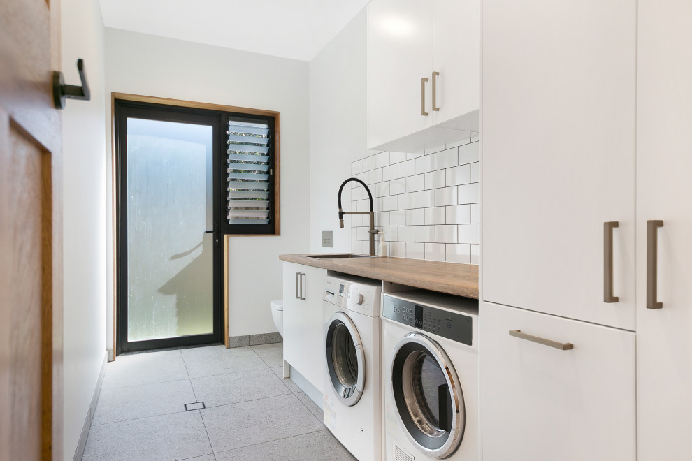 Photo of a contemporary single-wall dedicated laundry room in Sydney with a drop-in sink, flat-panel cabinets, white cabinets, wood benchtops, white splashback, subway tile splashback, ceramic floors, a side-by-side washer and dryer, white walls, grey floor and brown benchtop.