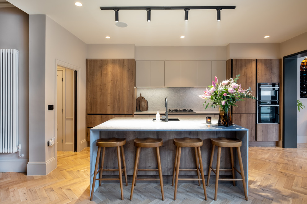 This is an example of a contemporary galley kitchen in Other with a submerged sink, flat-panel cabinets, medium wood cabinets, grey splashback, stone slab splashback, black appliances, medium hardwood flooring, an island, brown floors and beige worktops.