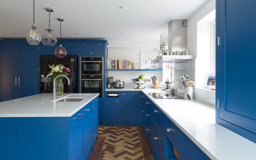 Design ideas for a mid-sized transitional l-shaped eat-in kitchen in Surrey with an integrated sink, shaker cabinets, blue cabinets, solid surface benchtops, white splashback, black appliances, medium hardwood floors and with island.