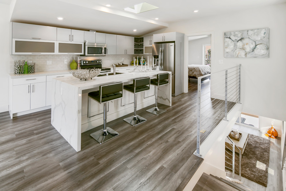 Design ideas for a mid-sized modern l-shaped open plan kitchen in San Francisco with a double-bowl sink, flat-panel cabinets, white cabinets, marble benchtops, white splashback, ceramic splashback, stainless steel appliances, with island and laminate floors.