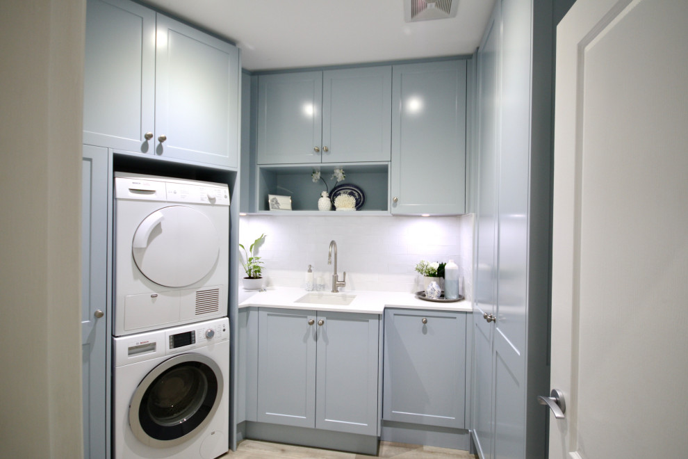 Design ideas for a mid-sized transitional u-shaped dedicated laundry room in Sydney with an undermount sink, shaker cabinets, blue cabinets, quartz benchtops, white splashback, subway tile splashback, white walls, light hardwood floors, a stacked washer and dryer, brown floor and white benchtop.