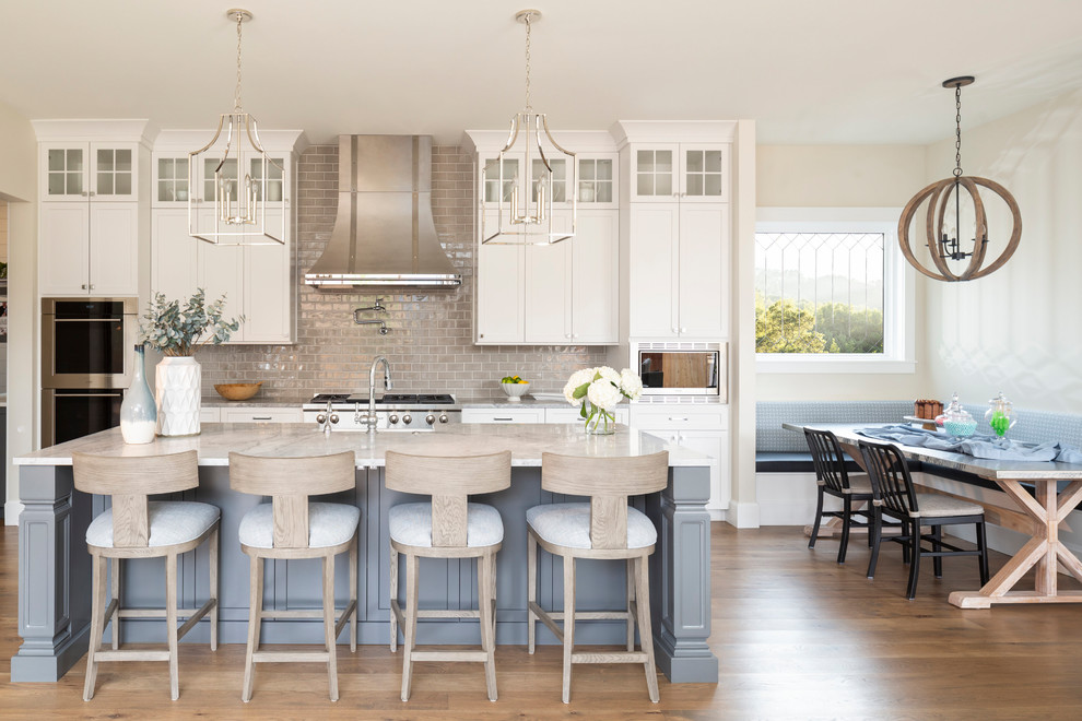 Design ideas for a beach style eat-in kitchen in Denver with shaker cabinets, white cabinets, grey splashback, subway tile splashback, stainless steel appliances, with island, grey benchtop, marble benchtops and light hardwood floors.