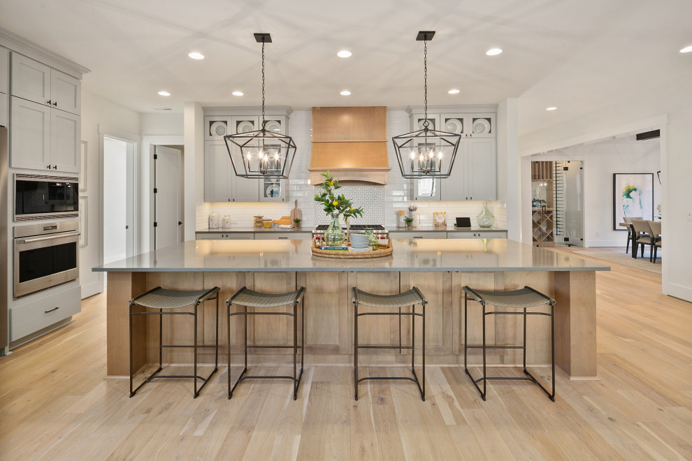 Large country l-shaped kitchen/diner in Seattle with a belfast sink, recessed-panel cabinets, grey cabinets, engineered stone countertops, white splashback, stainless steel appliances, light hardwood flooring, an island, beige floors and grey worktops.