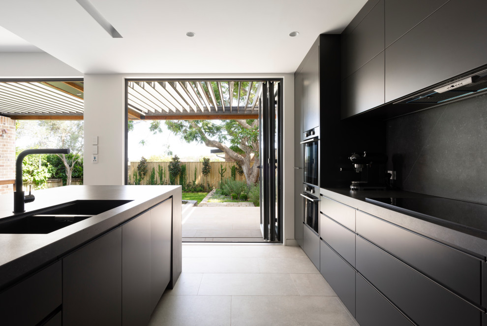 This is an example of a mid-sized contemporary galley open plan kitchen in Sydney with an undermount sink, grey cabinets, solid surface benchtops, grey splashback, stone slab splashback, black appliances, cement tiles, with island, grey floor and grey benchtop.