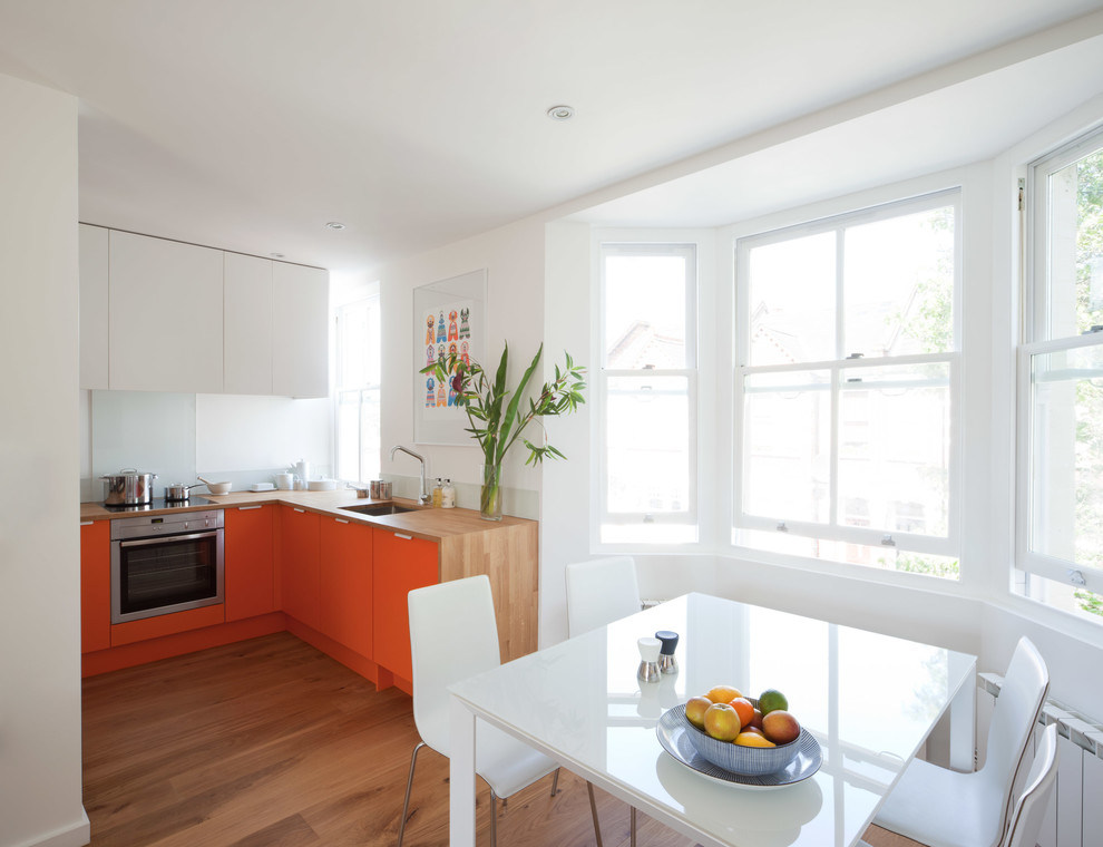 Small contemporary l-shaped open plan kitchen in London with an undermount sink, flat-panel cabinets, orange cabinets, wood benchtops, white splashback, glass sheet splashback, stainless steel appliances, light hardwood floors and no island.