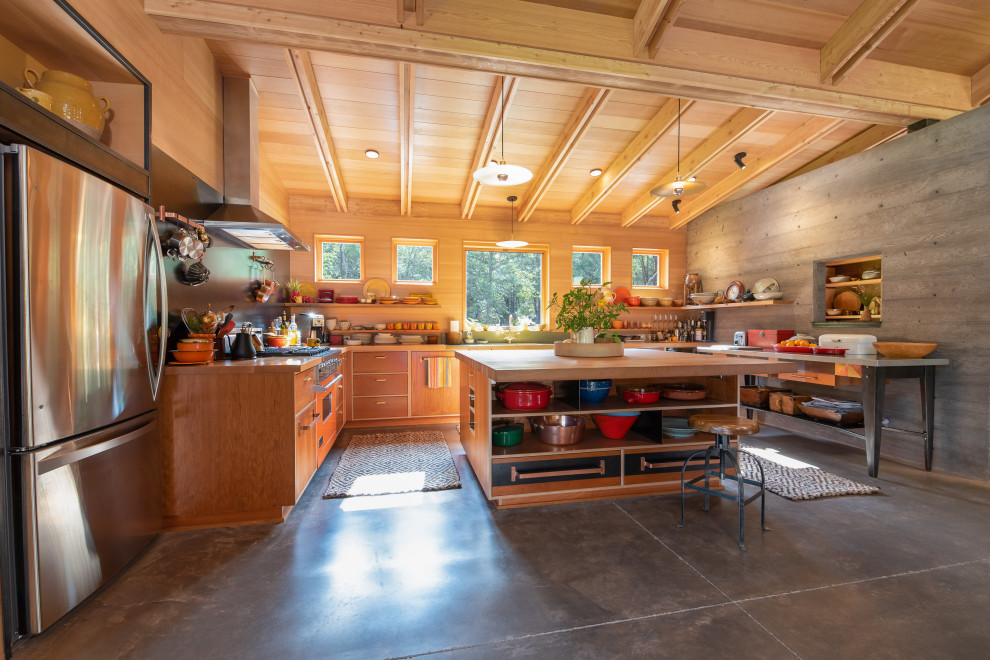This is an example of a country u-shaped open plan kitchen in Other with medium wood cabinets, wood benchtops, stainless steel appliances, concrete floors, with island, grey floor, flat-panel cabinets and brown benchtop.
