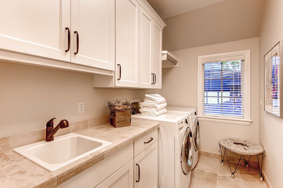 This is an example of a mid-sized transitional single-wall dedicated laundry room in Denver with a drop-in sink, shaker cabinets, white cabinets, tile benchtops, grey walls, porcelain floors and a side-by-side washer and dryer.