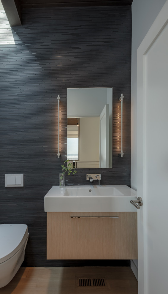 Mid-sized contemporary powder room in San Francisco with flat-panel cabinets, light wood cabinets, a wall-mount toilet, black tile, black walls, an integrated sink, solid surface benchtops, beige floor, white benchtops, light hardwood floors and mosaic tile.