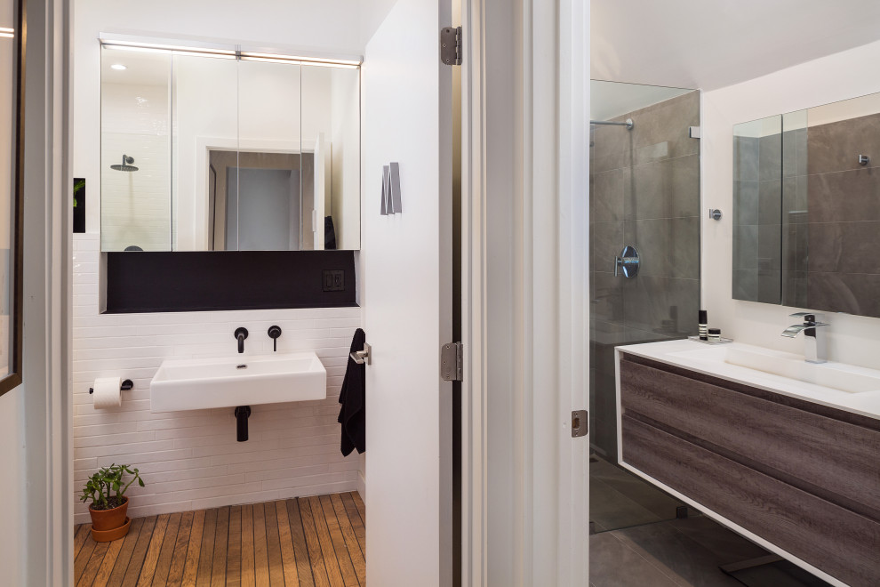 Design ideas for a mid-sized contemporary 3/4 wet room bathroom in Los Angeles with flat-panel cabinets, medium wood cabinets, a wall-mount toilet, gray tile, porcelain tile, white walls, porcelain floors, a wall-mount sink, solid surface benchtops, brown floor, an open shower and white benchtops.