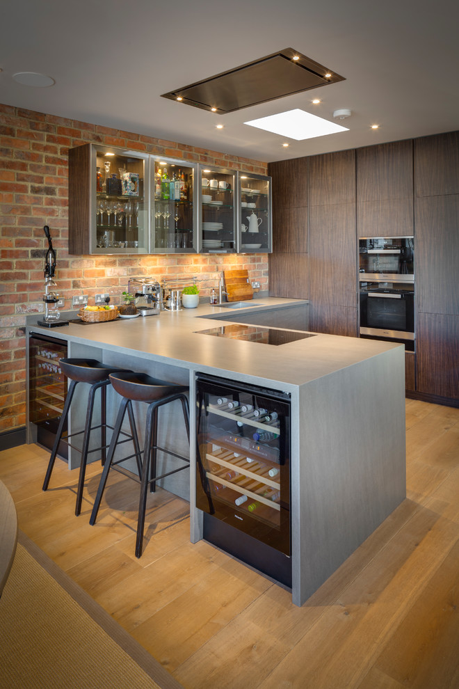 This is an example of a mid-sized contemporary u-shaped kitchen in London with flat-panel cabinets, brick splashback, stainless steel appliances, light hardwood floors, a peninsula, beige floor and grey benchtop.