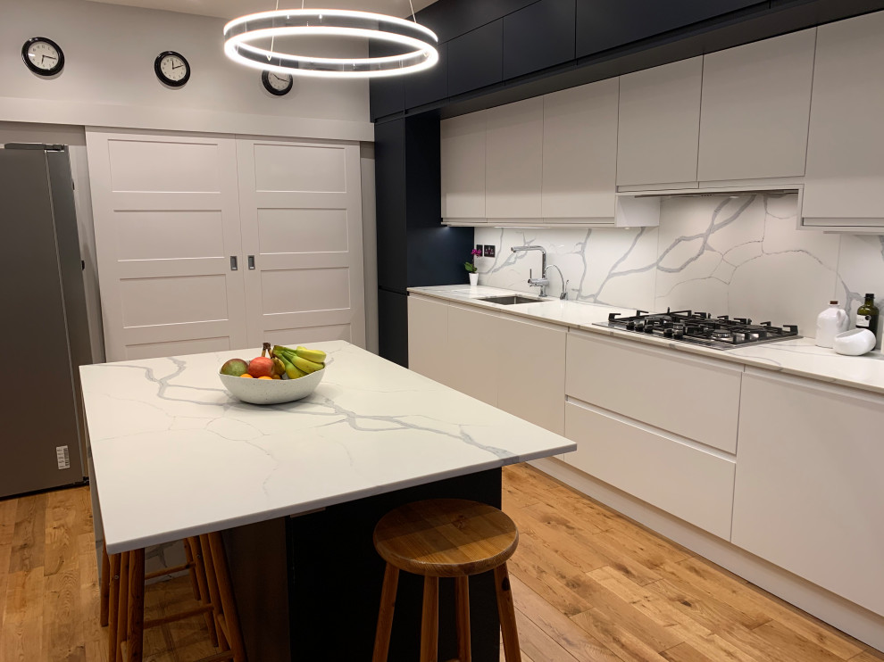 Modern open plan kitchen in London with flat-panel cabinets, blue cabinets, quartzite benchtops, white splashback, panelled appliances, medium hardwood floors, with island and white benchtop.