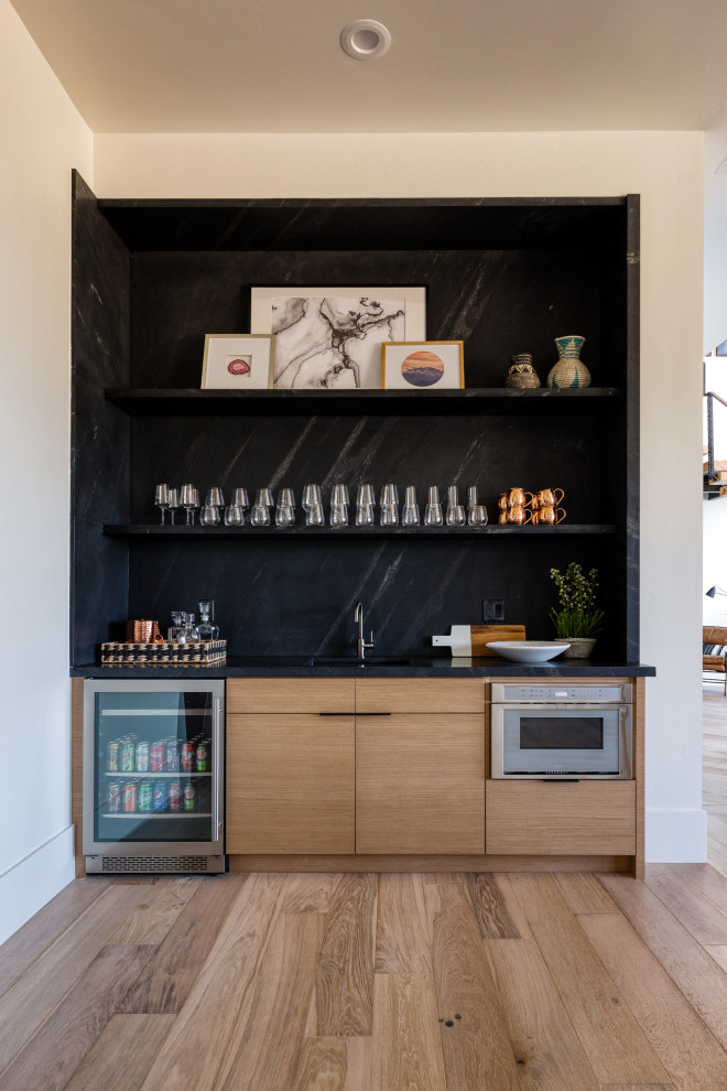 This is an example of a country single-wall wet bar in Salt Lake City with an undermount sink, flat-panel cabinets, light wood cabinets, black splashback, light hardwood floors, beige floor and black benchtop.