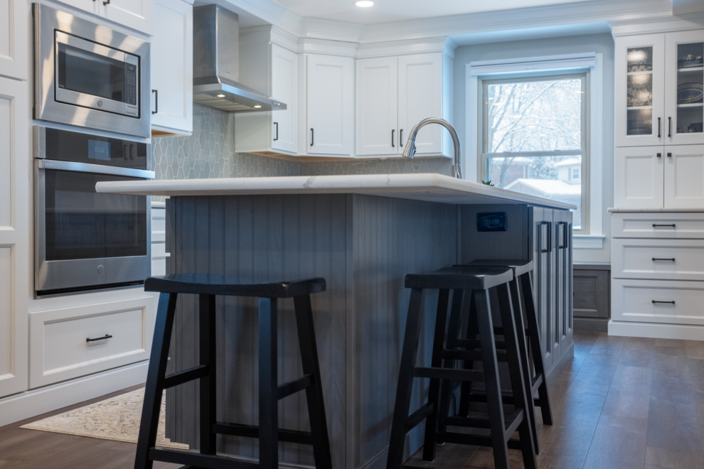 Large traditional single-wall eat-in kitchen in Cincinnati with a single-bowl sink, raised-panel cabinets, white cabinets, granite benchtops, grey splashback, porcelain splashback, stainless steel appliances, dark hardwood floors, with island, brown floor and white benchtop.