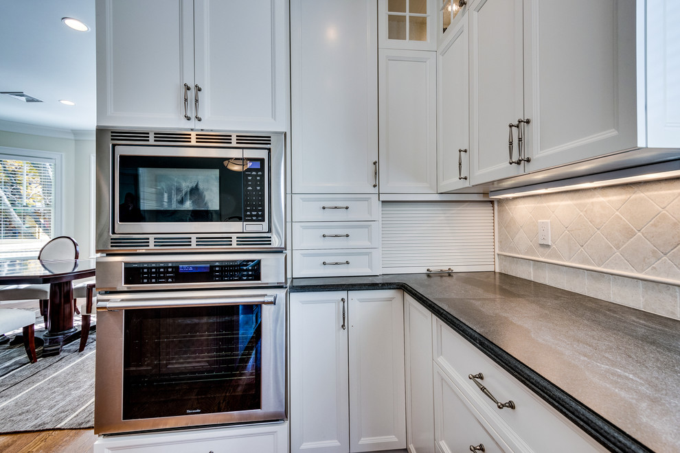 Open concept kitchen - mid-sized transitional l-shaped medium tone wood floor and brown floor open concept kitchen idea in Boston with a farmhouse sink, recessed-panel cabinets, white cabinets, soapstone countertops, beige backsplash, ceramic backsplash, stainless steel appliances and an island