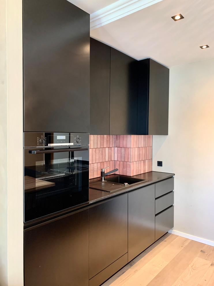 Photo of a mid-sized contemporary galley eat-in kitchen in Berlin with a drop-in sink, flat-panel cabinets, black cabinets, granite benchtops, pink splashback, ceramic splashback, black appliances, light hardwood floors, with island and black benchtop.