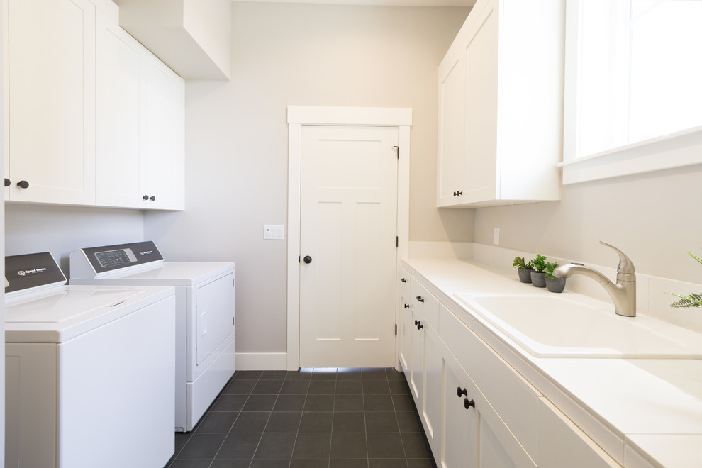 Mid-sized country galley dedicated laundry room in Portland with a drop-in sink, shaker cabinets, grey walls, a side-by-side washer and dryer, black floor and white benchtop.