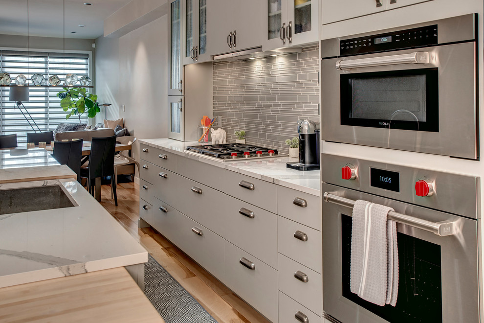 Example of a mid-sized minimalist light wood floor and beige floor eat-in kitchen design in Seattle with an undermount sink, glass-front cabinets, gray cabinets, quartz countertops, gray backsplash, glass tile backsplash, stainless steel appliances, an island and white countertops