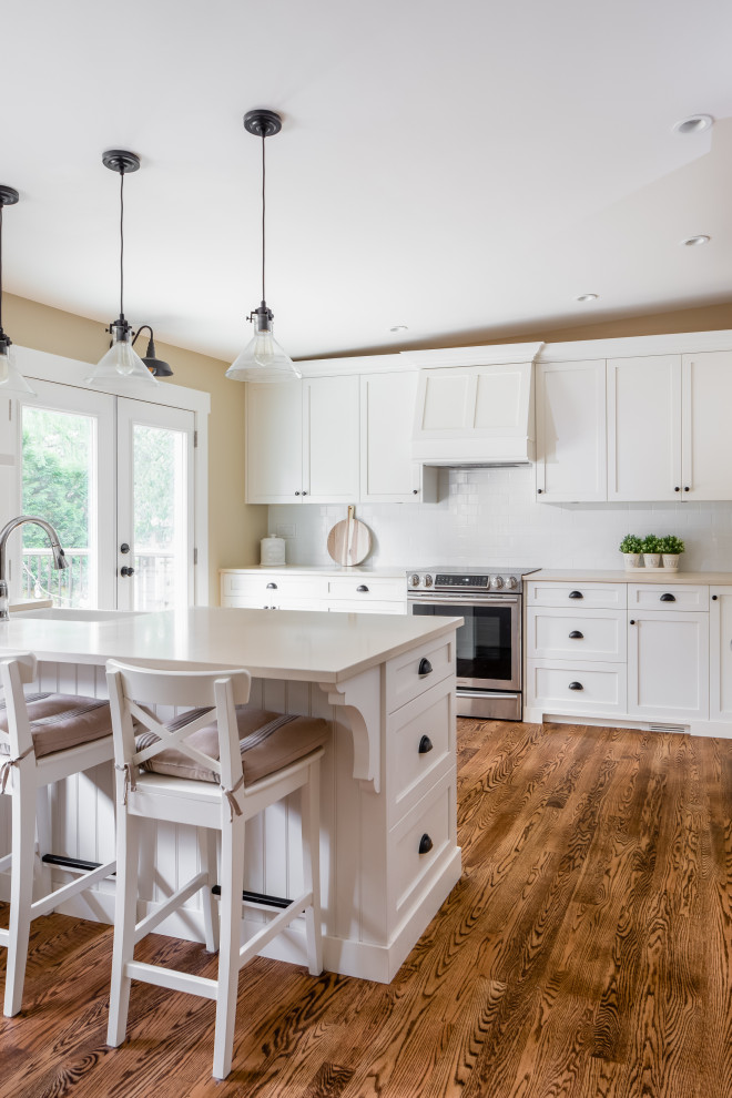 Example of a large mountain style l-shaped medium tone wood floor and brown floor eat-in kitchen design in Vancouver with a farmhouse sink, shaker cabinets, white cabinets, quartzite countertops, white backsplash, subway tile backsplash, stainless steel appliances, an island and white countertops