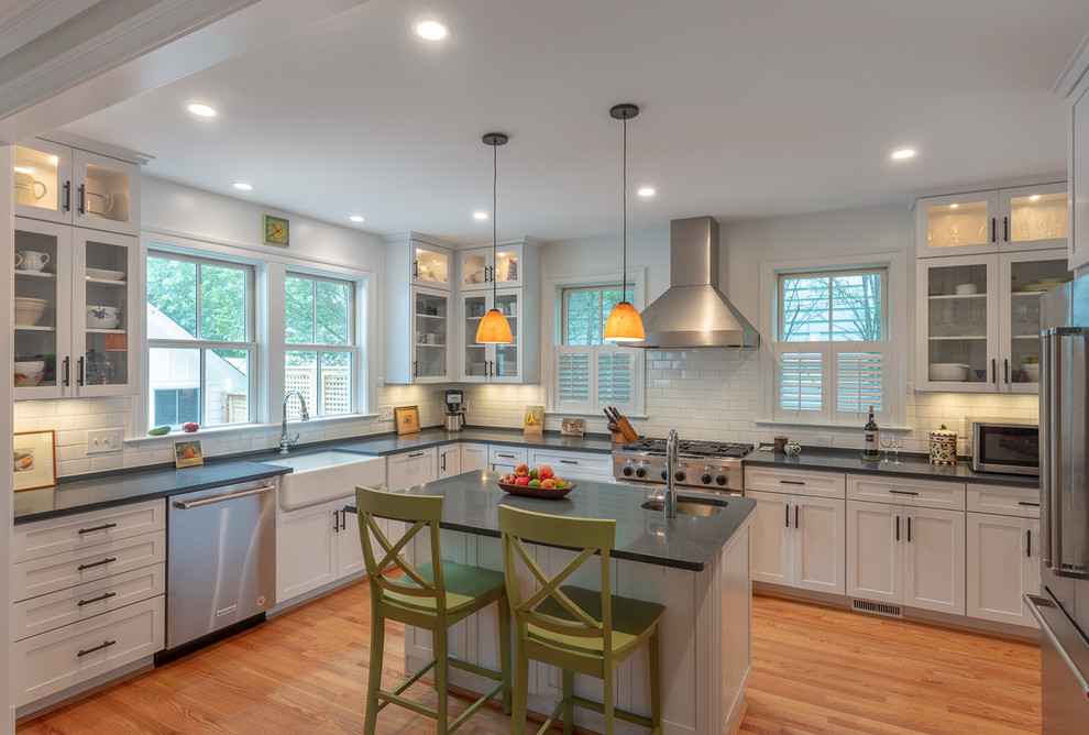 This is an example of a country u-shaped separate kitchen in DC Metro with a farmhouse sink, shaker cabinets, white cabinets, white splashback, subway tile splashback, stainless steel appliances, medium hardwood floors, with island, brown floor and black benchtop.
