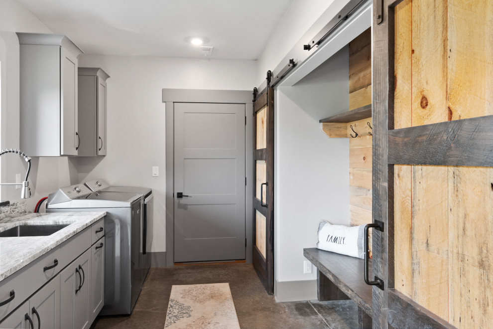 Photo of a large country galley dedicated laundry room in Milwaukee with an undermount sink, shaker cabinets, grey cabinets, quartz benchtops, multi-coloured splashback, engineered quartz splashback, white walls, concrete floors, a side-by-side washer and dryer, brown floor and multi-coloured benchtop.